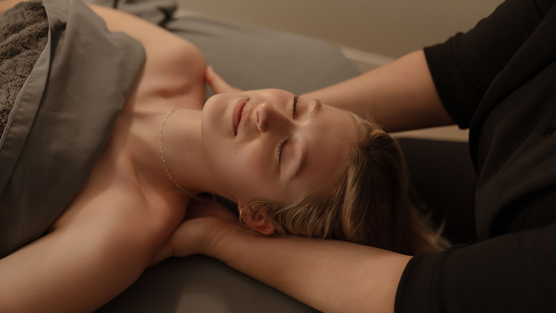 A serene scene of a woman lying on her back during a relaxation massage, with a practitioner's hands gently supporting her neck, in a dimly lit room with muted colors, conveying a sense of calm and tranquility.