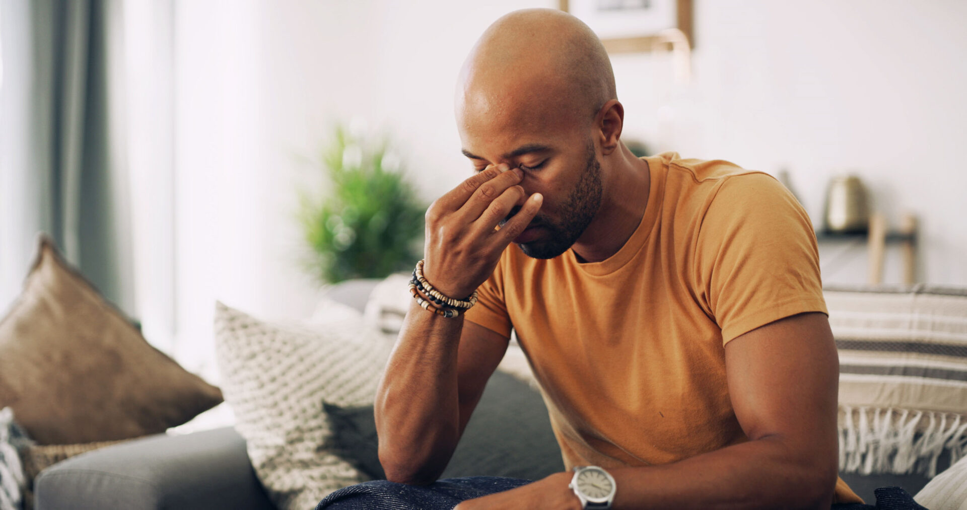 A man suffering with a headache and sinus pressure from overworking and seasonal change, sitting on a couch.