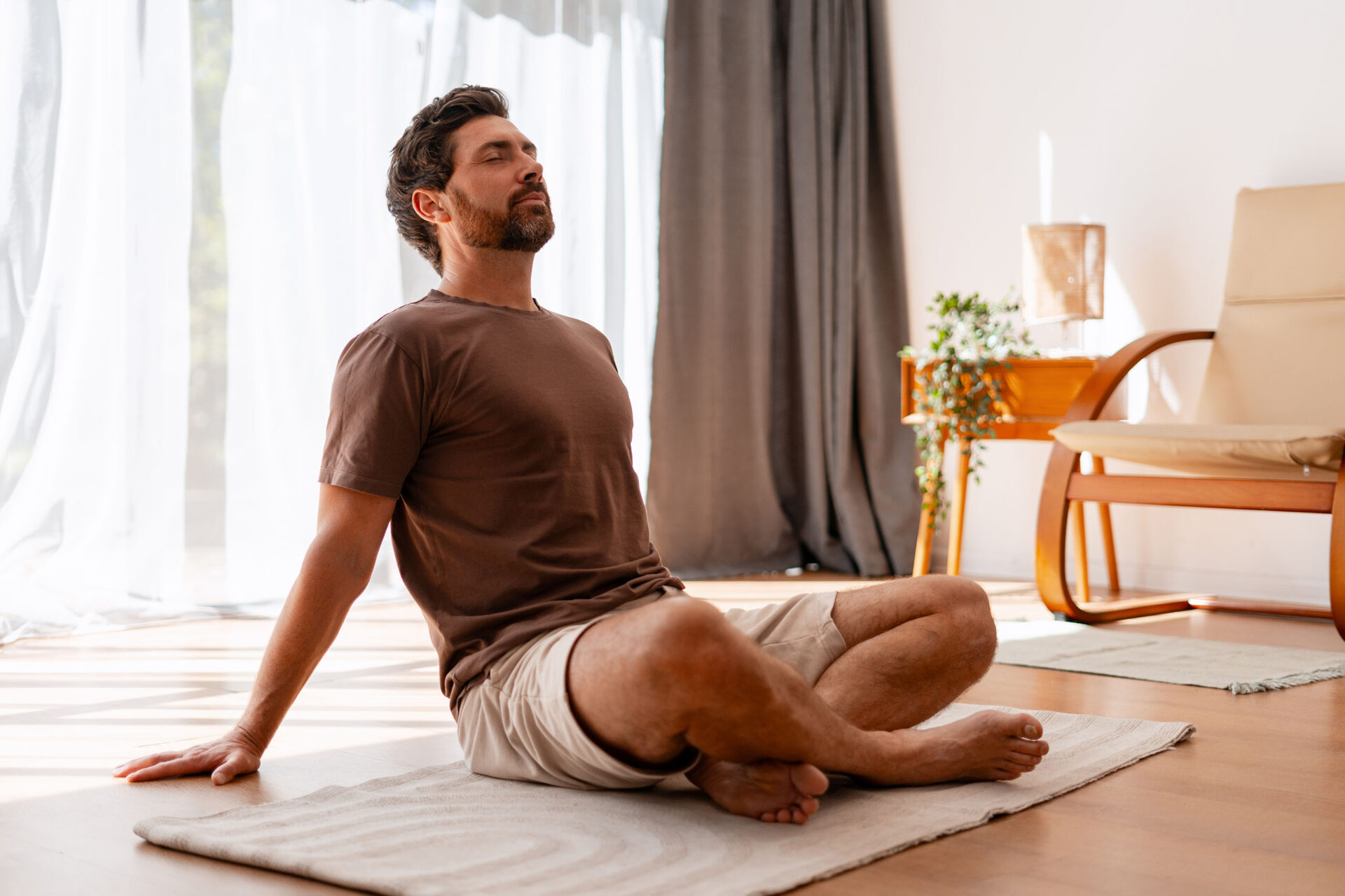 Calm man sitting cross-legged on mat in living room, practicing mindfulness and enjoying a peaceful moment of relaxation.