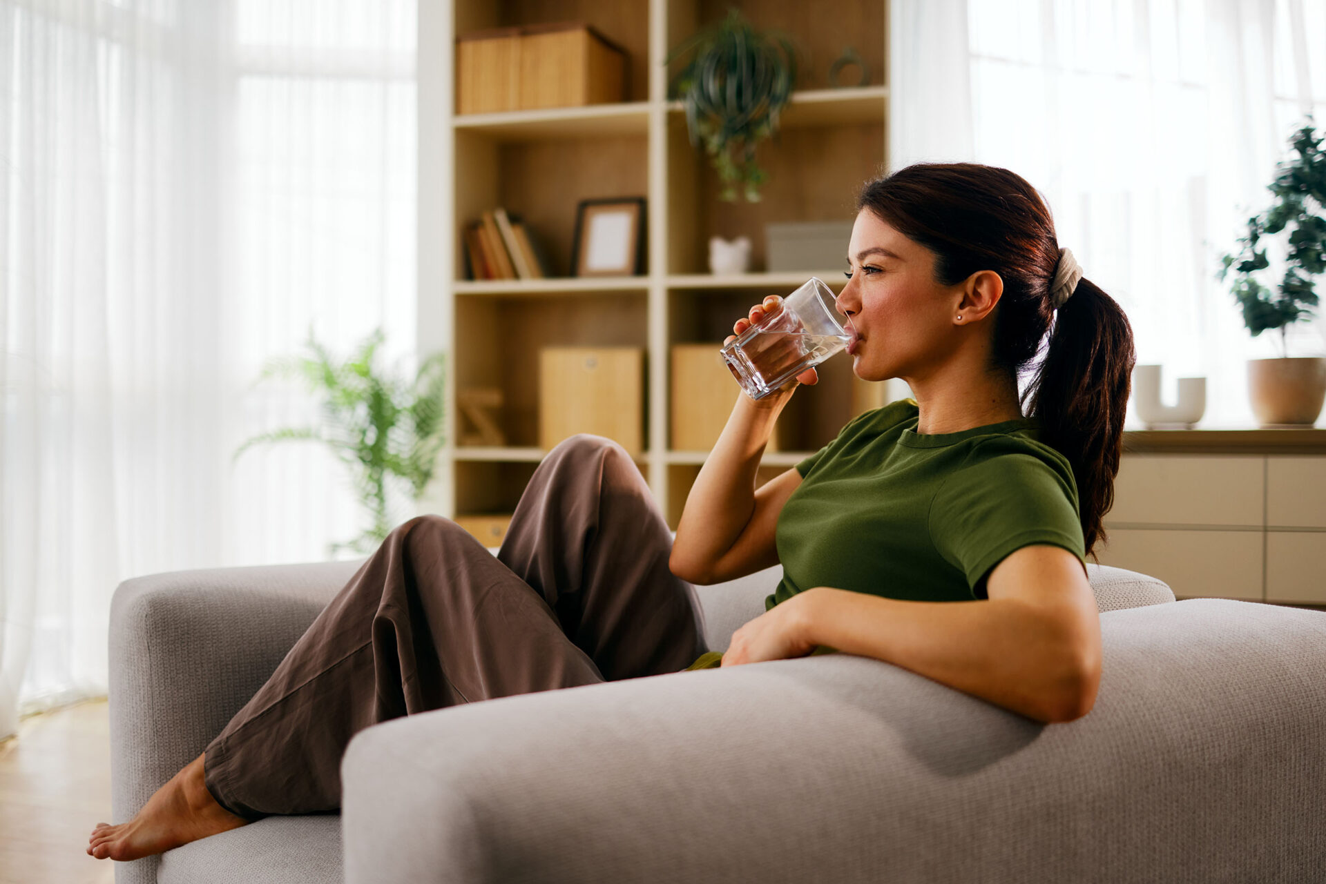 A woman lounging on a couch, drinking a glass of water after feeling completely relaxed from her wellness therapies.